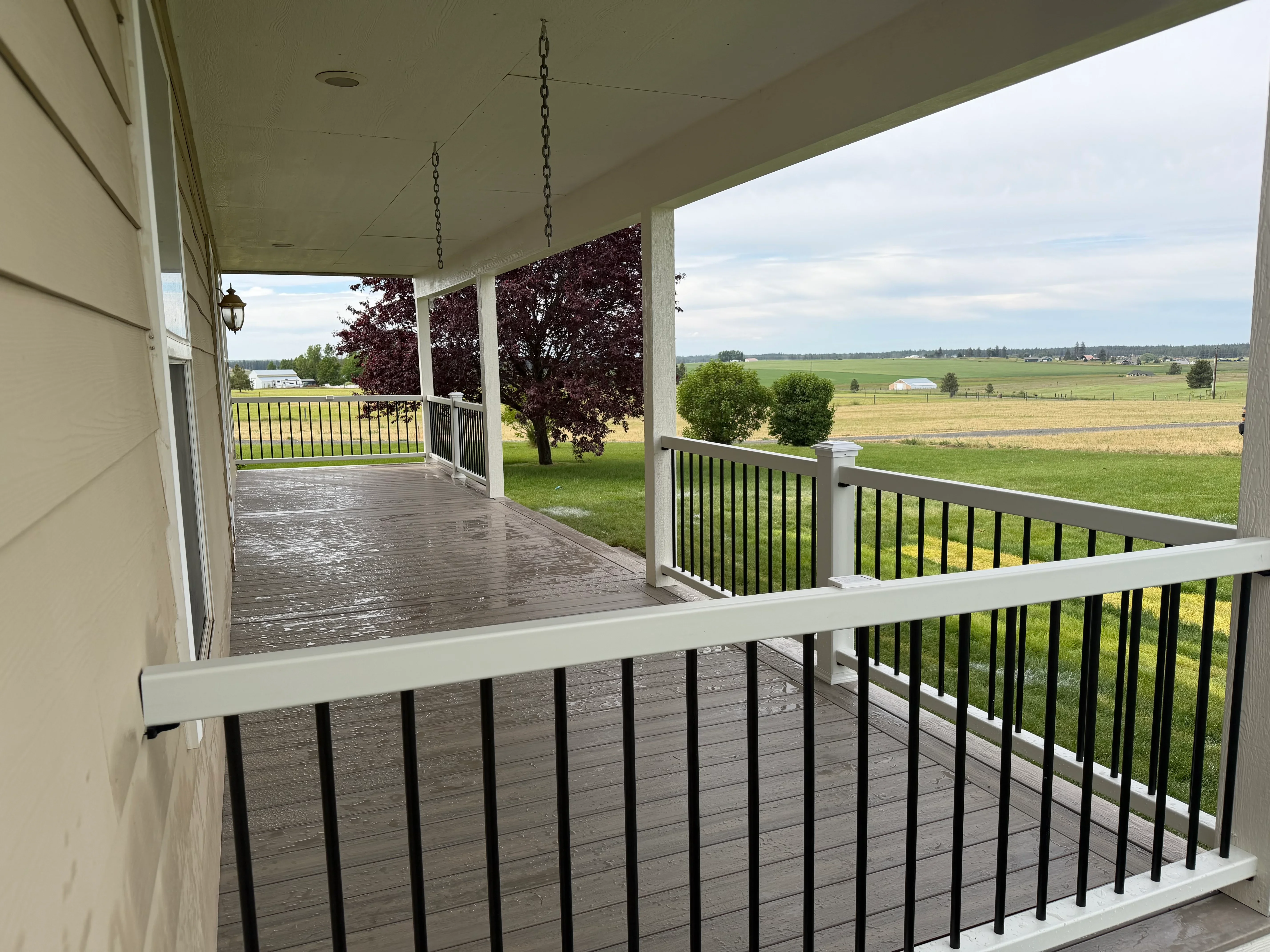 Covered porch with composite decking, white posts, and black metal railing overlooking rural farmland